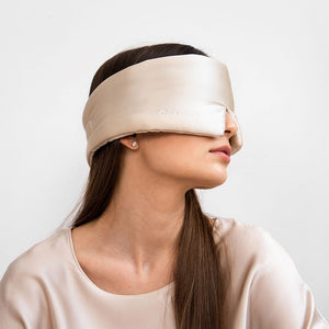 An older person with long gray hair wears a luxurious beige DROWSY - SATIN SLEEP MASK by DROWSY SLEEP CO, made of mulberry silk. In a matching satin top, they stand against a plain white background, ready for beauty sleep.