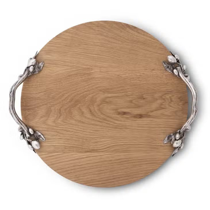 The OLIVE GROVE CHEESE BOARD by VAGABOND HOUSE is a round oak wood serving tray with a smooth surface and two ornately designed pewter-embellished handles on opposite sides. The handles feature intricate leaf and berry patterns, adding an elegant touch to the rustic tray.