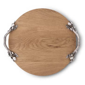 The OLIVE GROVE CHEESE BOARD by VAGABOND HOUSE is a round oak wood serving tray with a smooth surface and two ornately designed pewter-embellished handles on opposite sides. The handles feature intricate leaf and berry patterns, adding an elegant touch to the rustic tray.