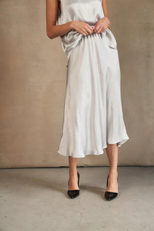 A woman models the ROSE & CROWN - SATIN MIDI SKIRT in silky blue, paired with a short-sleeved light blue knit top, standing against a white background. Her head is not visible in the image.