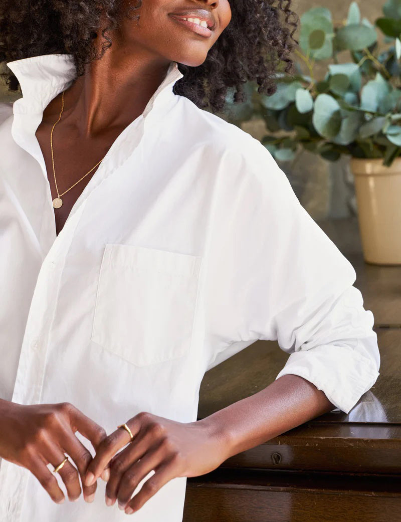 A person with curly hair, dressed in the FRANK & EILEEN OONA Continuous Sleeve Button Up Shirt in Superluxe Cotton White, and relaxed-fit green pants, stands facing away from the camera in front of a piano with their hands clasped behind their back. A stone wall and plants provide the backdrop.
