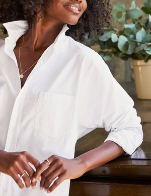 A person with curly hair, dressed in the FRANK & EILEEN OONA Continuous Sleeve Button Up Shirt in Superluxe Cotton White, and relaxed-fit green pants, stands facing away from the camera in front of a piano with their hands clasped behind their back. A stone wall and plants provide the backdrop.