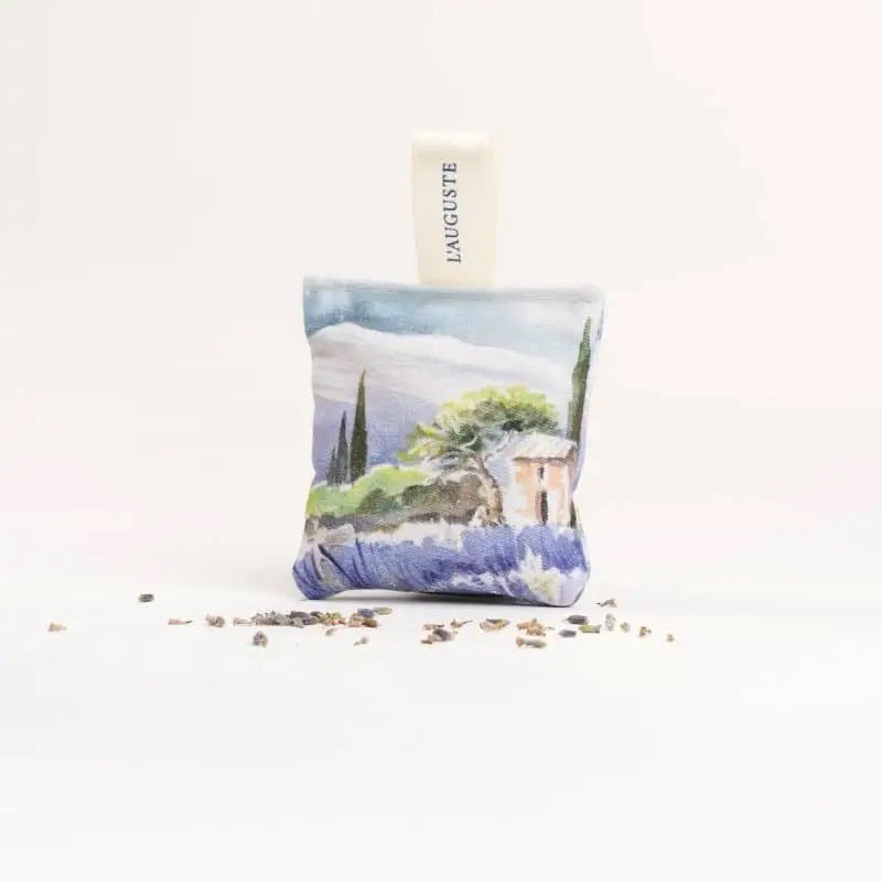A sachet from L&#39;AUGUSTE PROVENCE, featuring a watercolor landscape and &quot;Provence&quot; fabric tag, stands upright on a white surface amid dried lavender. This French artisanal gift comes in a FRENCH ORGANIC LAVENDER SACHET AND GIFT BOX.