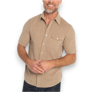 A man wearing the CRIQUET Short Sleeve Corduroy Pearl Snap Shirt in Irish Coffee stands in front of a dark door and red brick wall, holding a black object at chest level.