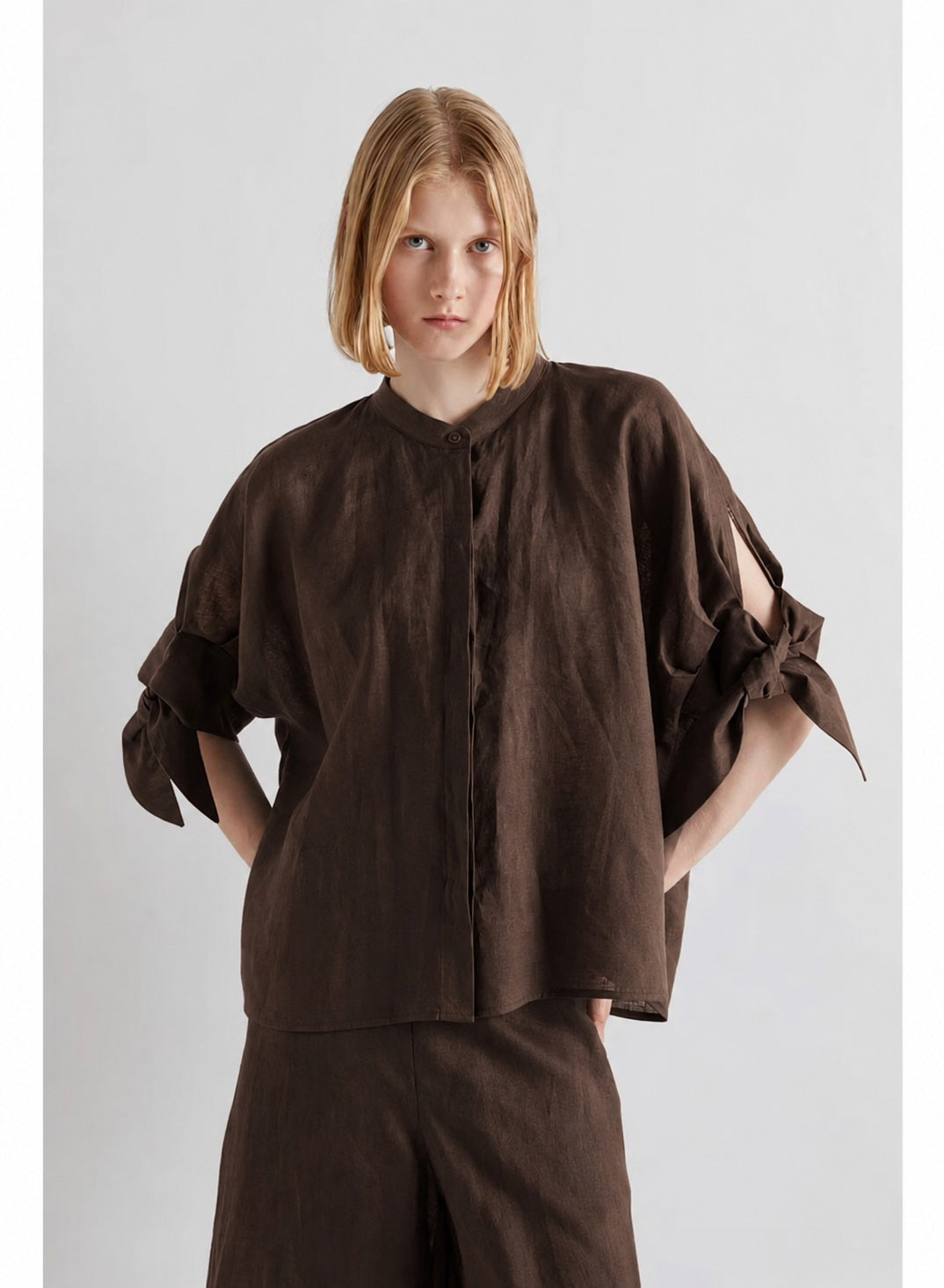 A woman with brown hair in a low ponytail is standing indoors, smiling at the camera while wearing the MEIMEIJ Linen Top with Tie Sleeve in Moka—a loose, buttoned blouse made of linen by MEIMEIJ, featuring short sleeves tied at the elbows and crafted in Italy for perfect summer style.