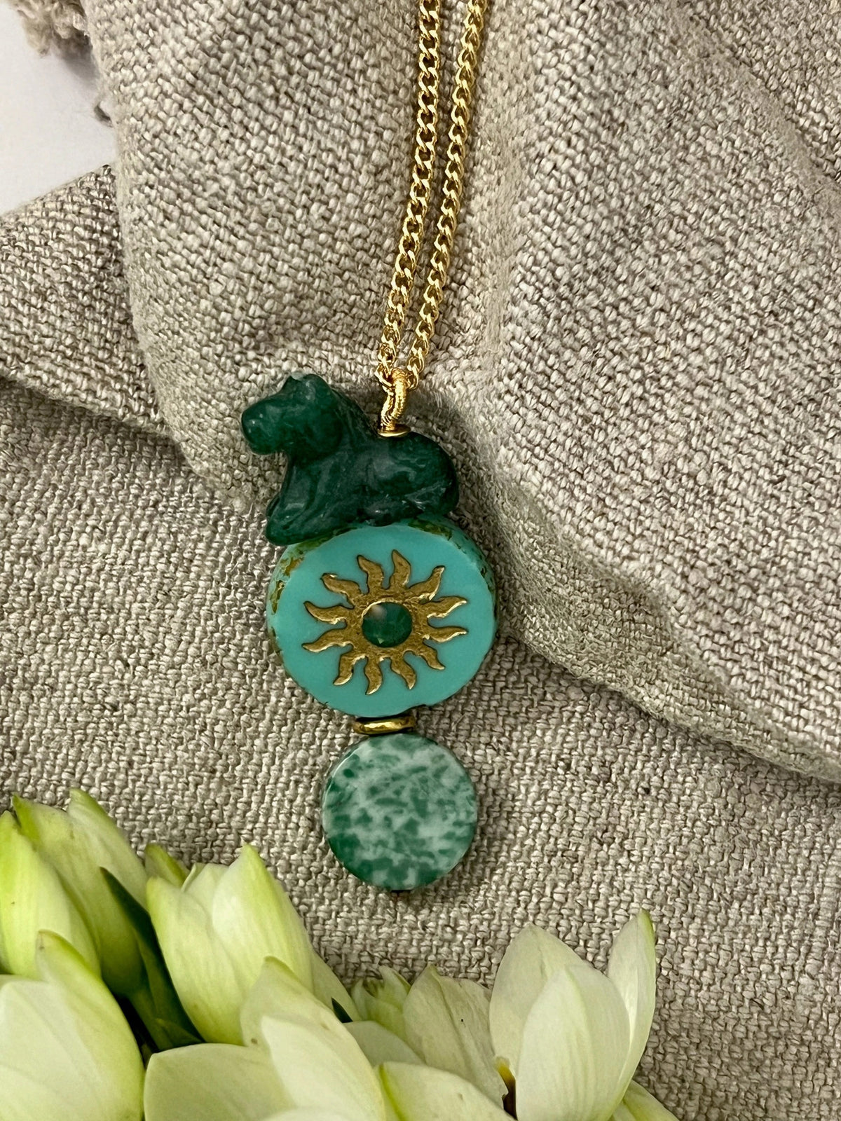 The SANDRINE FRANCE STUDIO - COMPANION AUSTIN NECKLACE by SANDRINE FRANCE STUDIO features a gold chain adorned with three green pendants: a small abstract shape, a jade round disk accented with a gold sun motif, and an oval bead with a marbled pattern—each detail showcasing the artistry of handmade NYC craftsmanship.
