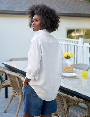 A person with curly hair wears the FRANK & EILEEN EILEEN Relaxed Button Up in Washed Linen White, standing outdoors against a blurred background of greenery and a building.