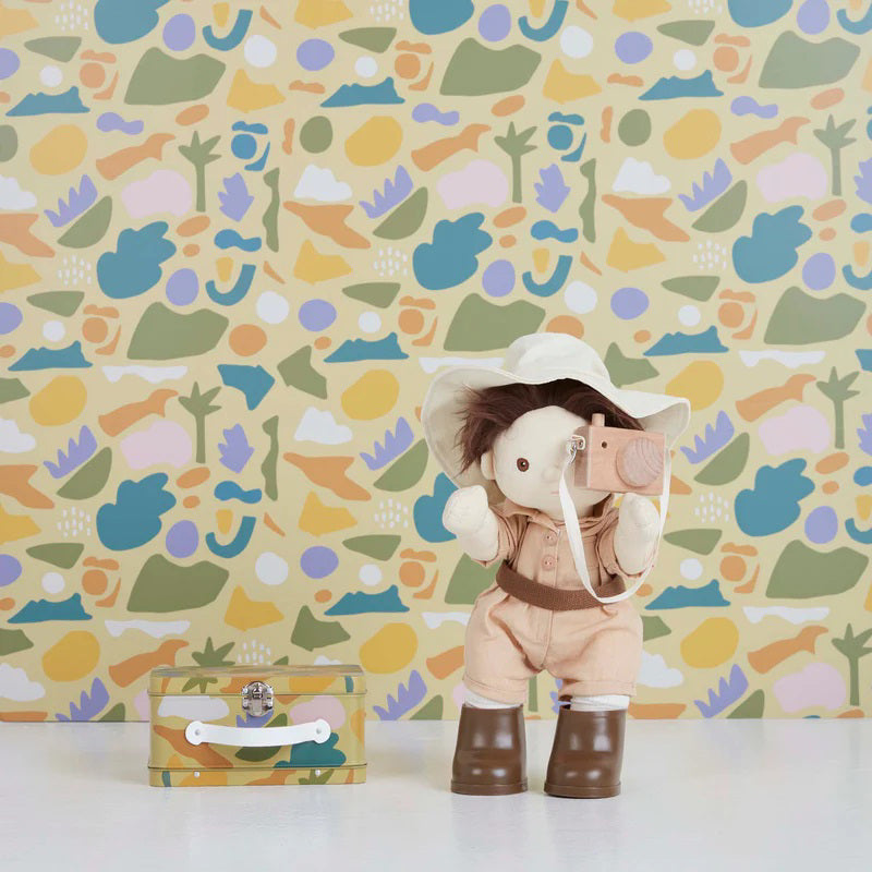 An OLLI ELLA USA DINKUM DOLL from the Pretend Pack Travel Sets, outfitted in a wide-brimmed hat, khaki overalls, and brown boots, poses with a petite suitcase. The backdrop showcases a vibrant, abstract pattern filled with diverse shapes and pastel hues, perfect for sparking endless imaginative adventures.