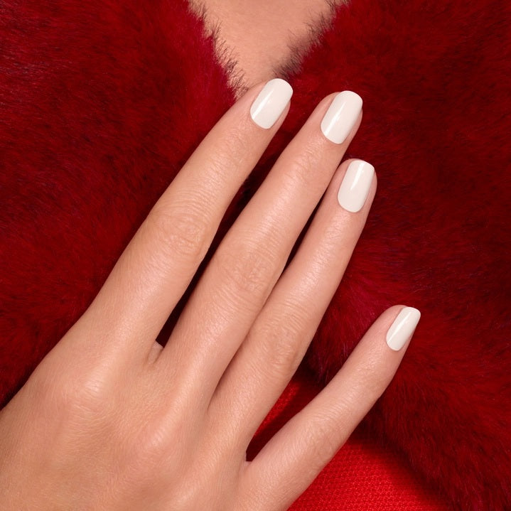 A hand with neatly manicured off-white nails, painted with MANUCURIST - GREEN™ NAIL POLISH IN CREAM by MANUCURIST INC, rests on a rich red textured fabric, creating a striking contrast between the nail color and the background.