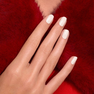 A hand with neatly manicured off-white nails, painted with MANUCURIST - GREEN™ NAIL POLISH IN CREAM by MANUCURIST INC, rests on a rich red textured fabric, creating a striking contrast between the nail color and the background.