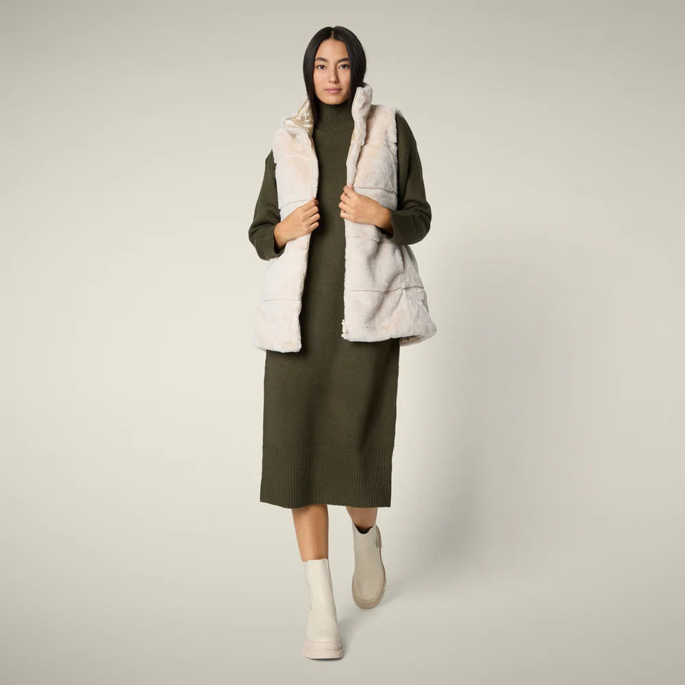 A woman with straight blonde hair wears the SAVE THE DUCK NIVES REVERSIBLE VEST over a white long-sleeve top, beige wide-leg pants, and black shoes, standing against a plain background and highlighting the vest&#39;s chic style.