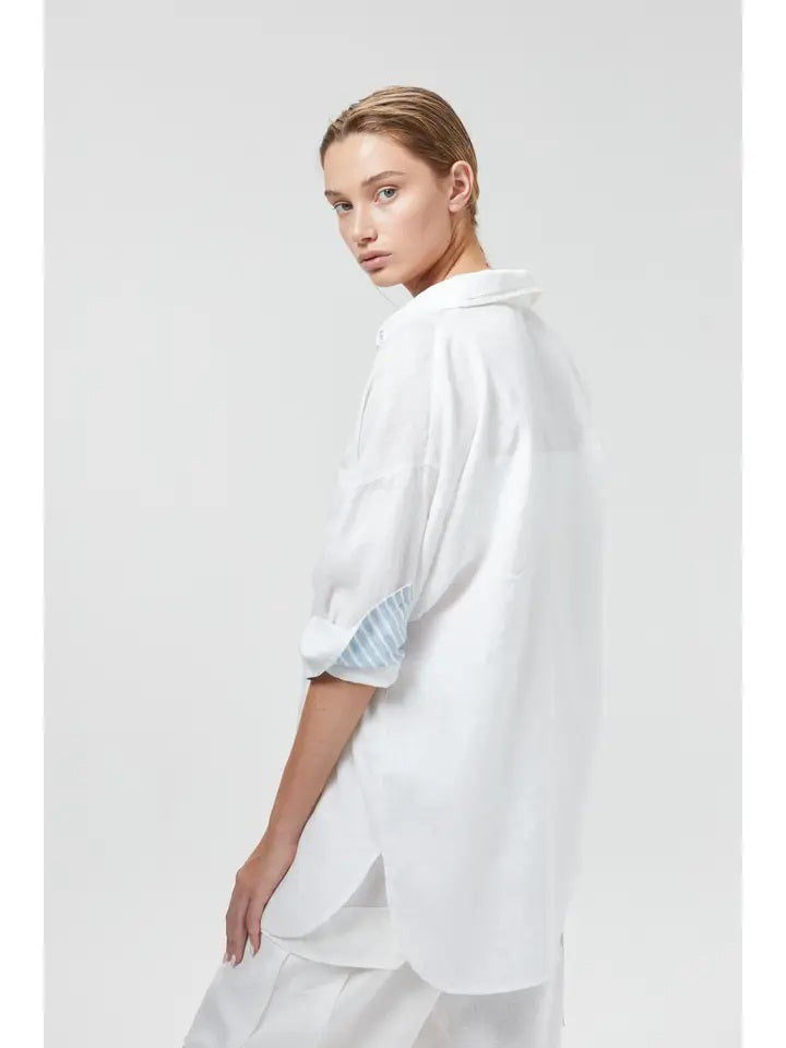 A person stands against a plain background, wearing the LANHTROPY - ISLAND LINEN OVERSIZED SHIRT from LANHTROPY, crafted from premium organic linen. The shirt features blue and white striped detailing on the cuffs and inner placket, paired with white pants. Elegant shell buttons accentuate the shirt. Their hands rest in the pockets of their pants as they gaze directly at the camera.