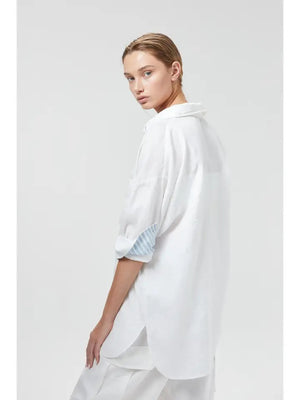 A person stands against a plain background, wearing the LANHTROPY - ISLAND LINEN OVERSIZED SHIRT from LANHTROPY, crafted from premium organic linen. The shirt features blue and white striped detailing on the cuffs and inner placket, paired with white pants. Elegant shell buttons accentuate the shirt. Their hands rest in the pockets of their pants as they gaze directly at the camera.