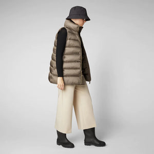 A person wearing a black cap, sunglasses, the SAVE THE DUCK - CORAL PUFFER VEST with animal-free down from SAVE THE DUCK over a black long-sleeve top, beige wide-legged pants, and black ankle boots stands against a plain background. The outfit appears to be styled for colder weather.