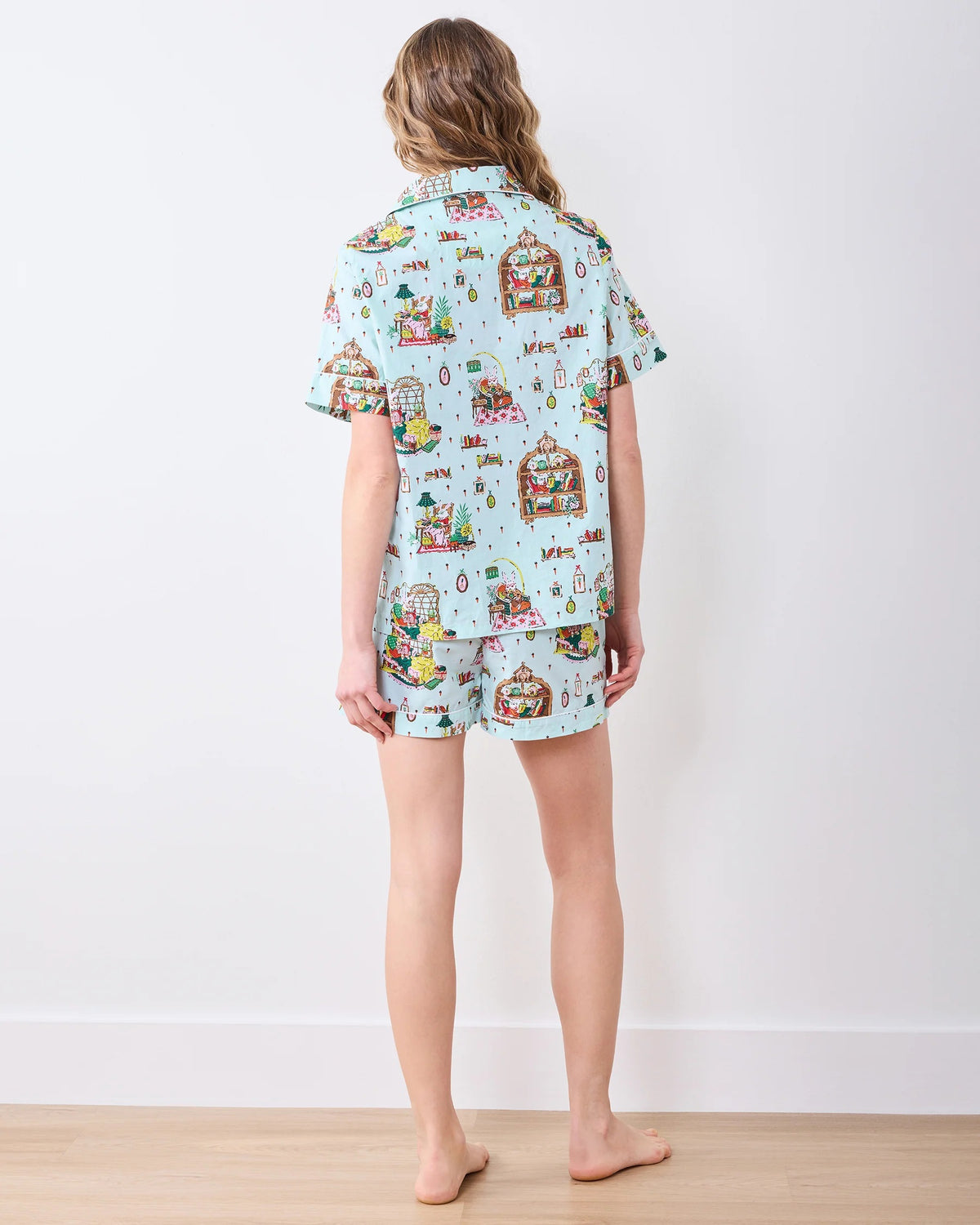 A person stands barefoot against a white wall, smiling and wearing the PRINTFRESH - READING NOOK PAJAMA SHORT SET by PRINTFRESH—colorful, organic cotton pajamas ideal for relaxing in your favorite reading nook.