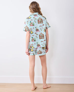 A person stands barefoot against a white wall, smiling and wearing the PRINTFRESH - READING NOOK PAJAMA SHORT SET by PRINTFRESH—colorful, organic cotton pajamas ideal for relaxing in your favorite reading nook.