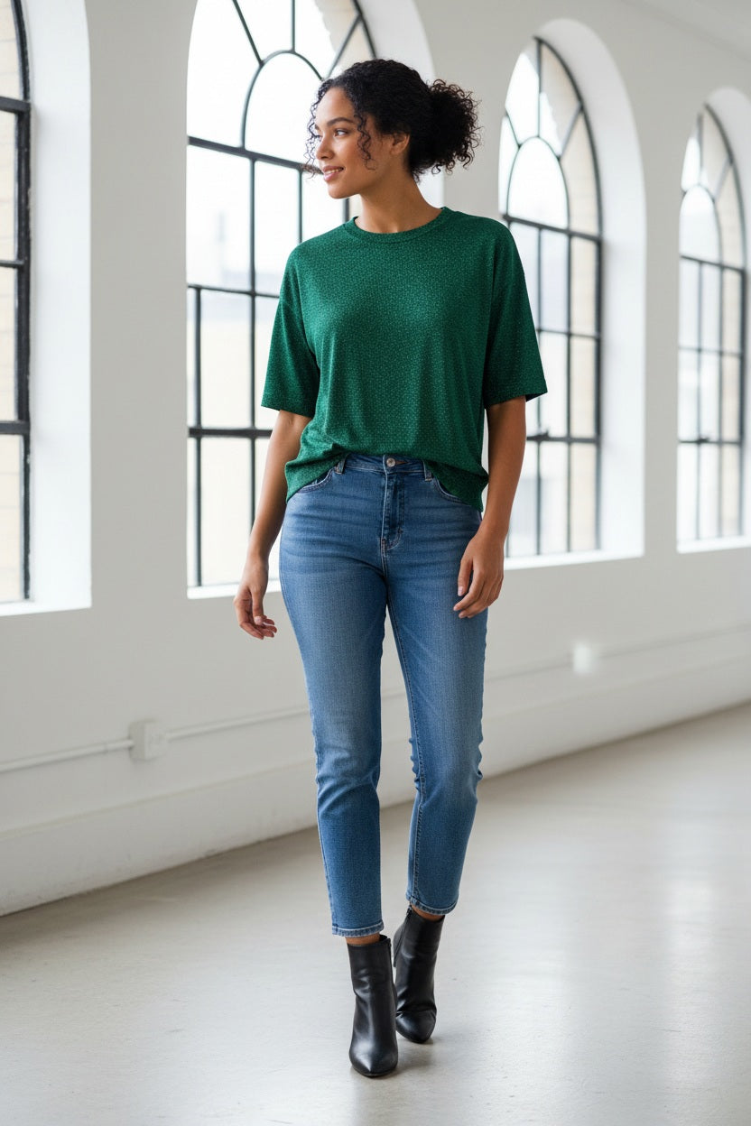A woman with long brown hair wears the MOMONI - IORA LUREX JERSEY T SHIRT in gold, paired with beige and blue wide-leg floral pants and brown sneakers, standing against a neutral studio background.