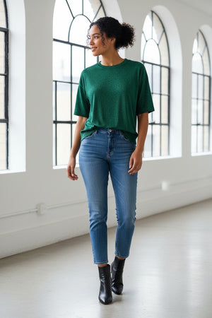 A woman with long brown hair wears the MOMONI - IORA LUREX JERSEY T SHIRT in gold, paired with beige and blue wide-leg floral pants and brown sneakers, standing against a neutral studio background.