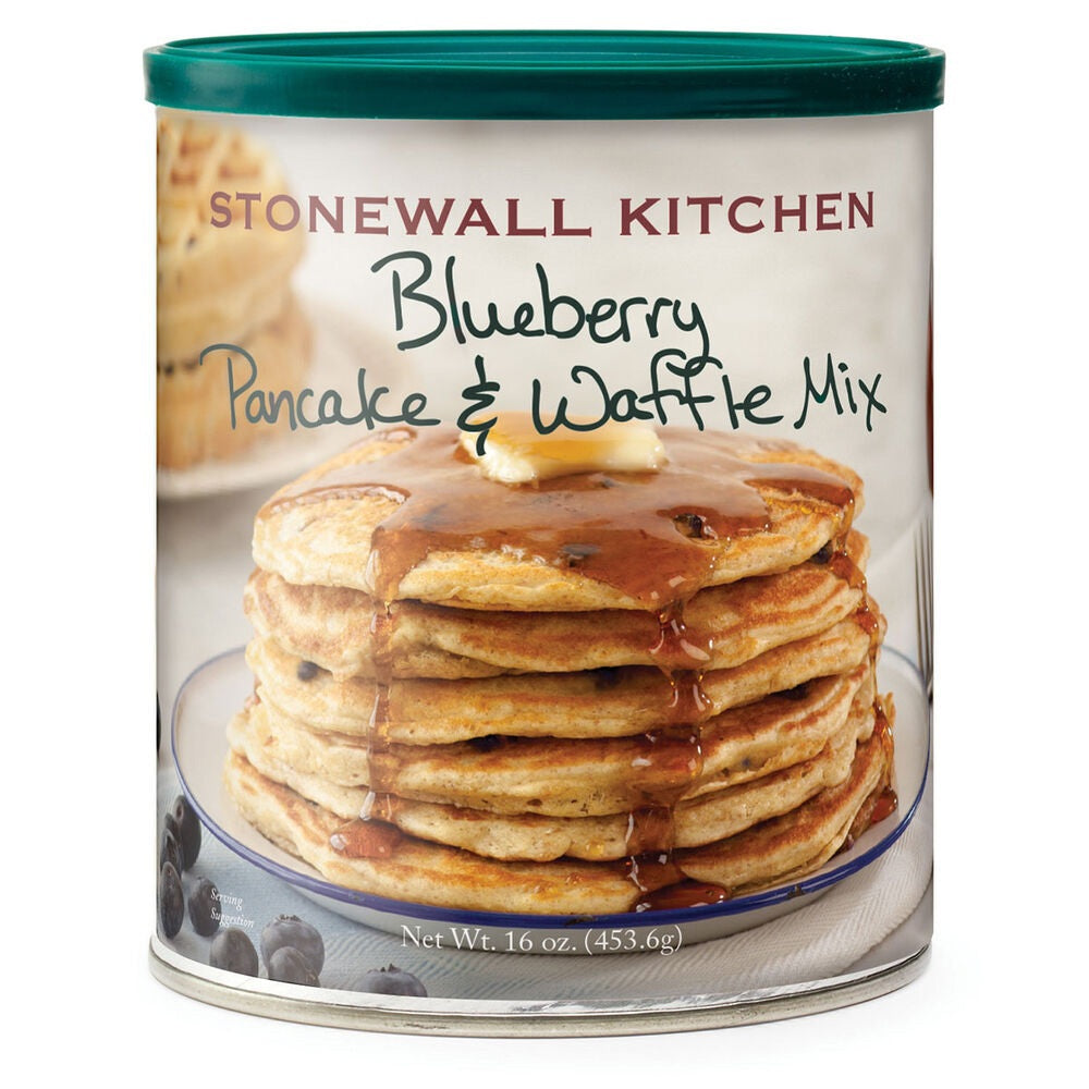 A stack of pancakes made with STONEWALL KITCHEN - BLUEBERRY PANCAKE AND WAFFLE MIX and topped with creamy butter sits alongside fresh Maine wild blueberries, a tempting pile of waffles, and a glass container of blueberry syrup in the background.