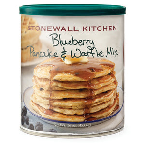 A stack of pancakes made with STONEWALL KITCHEN - BLUEBERRY PANCAKE AND WAFFLE MIX and topped with creamy butter sits alongside fresh Maine wild blueberries, a tempting pile of waffles, and a glass container of blueberry syrup in the background.