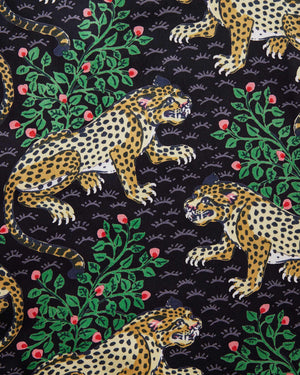 A woman stands barefoot on a woven rug in front of a white paneled wall, wearing the PRINTFRESH - Bagheera Satin Wildest Dream Pajama Set in Ink from PRINTFRESH, featuring a vibrant leopard and foliage pattern with wide-leg pants.
