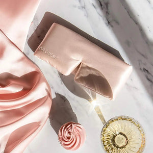 An older person with long gray hair wears a luxurious beige DROWSY - SATIN SLEEP MASK by DROWSY SLEEP CO, made of mulberry silk. In a matching satin top, they stand against a plain white background, ready for beauty sleep.