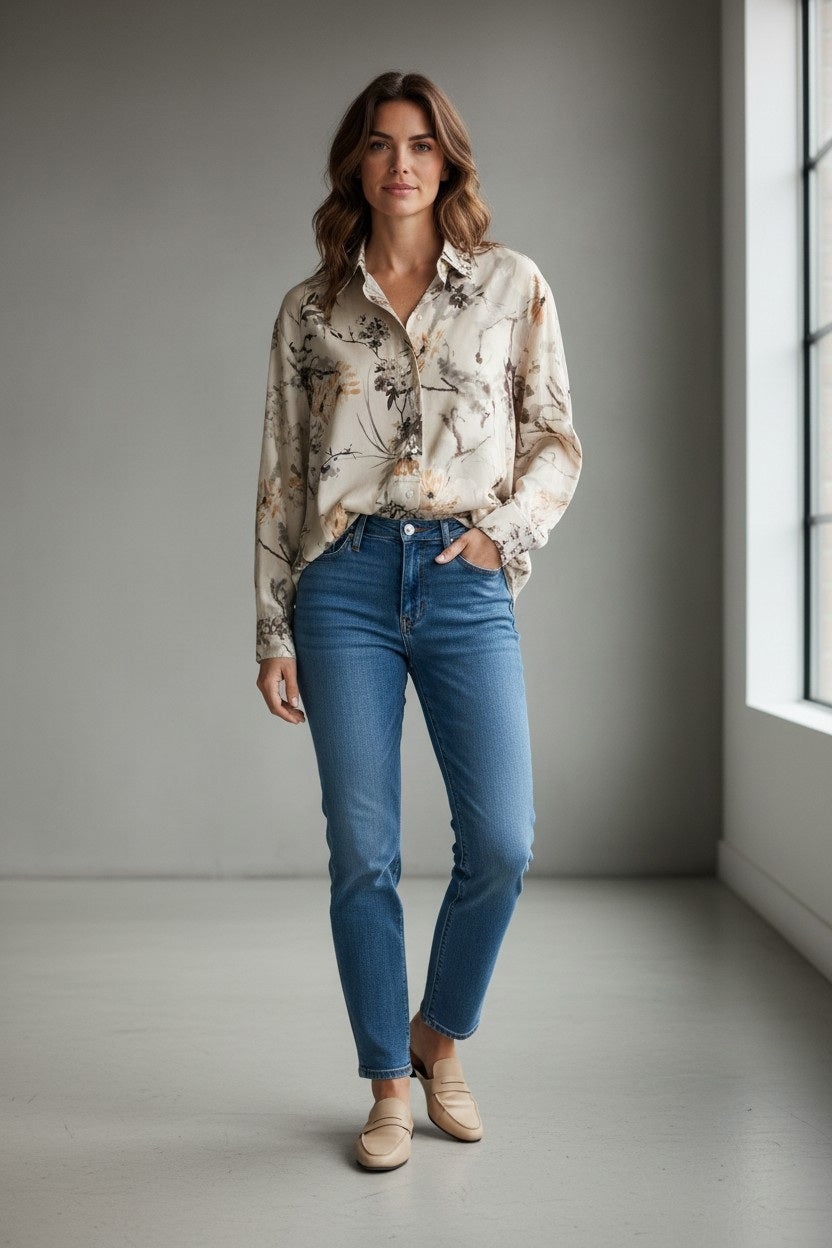 A woman stands indoors by a window, wearing the VLT’S BY VALENTINA - FLORAL VISCOSE BLOUSE from DANI CONFEZIONI, paired with blue jeans and beige loafers. She has one hand in her pocket and looks at the camera with a slight smile.