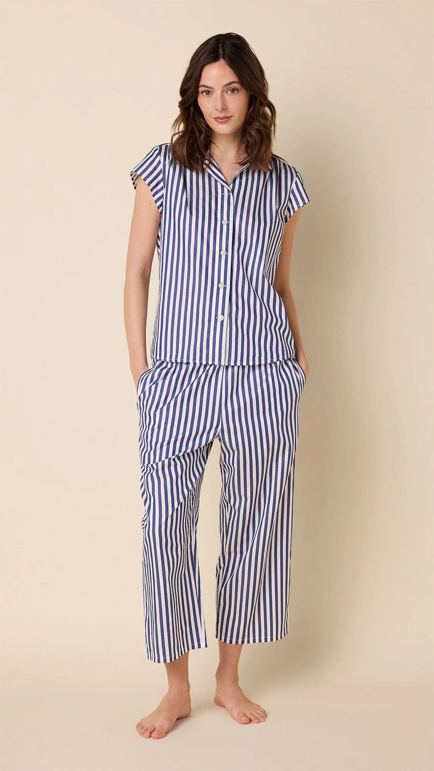 A woman wears CAT&#39;S PAJAMAS - CLASSIC STRIPE LUXE PIMA CAPRI PAJAMAS with blue and white vertical stripes, short sleeves, and cropped pants, standing barefoot against a plain beige background.