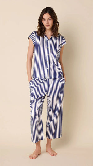 A woman wears CAT'S PAJAMAS - CLASSIC STRIPE LUXE PIMA CAPRI PAJAMAS with blue and white vertical stripes, short sleeves, and cropped pants, standing barefoot against a plain beige background.