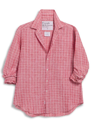 Wearing the FRANK & EILEEN Eileen Relaxed Button Up in Italian Linen Red and White Grid, a woman with blue jeans and white sneakers stands smiling at an outdoor cafe surrounded by wooden chairs and lush green plants.
