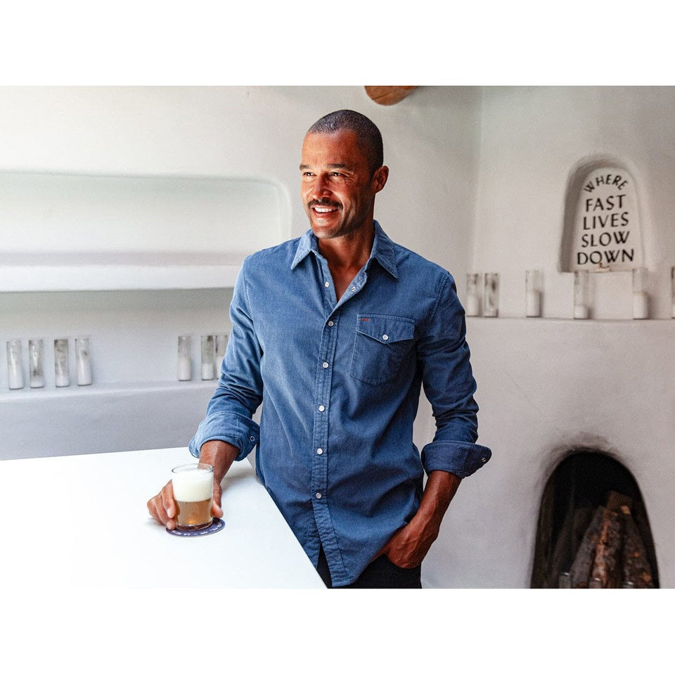 A man is wearing the CRIQUET Long Sleeve Corduroy Pearl Snap Shirt in Slate Blue by CRIQUET, featuring a chest pocket and rolled-up cuffs. He pairs it with brown pants against a white background.
