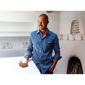 A man is wearing the CRIQUET Long Sleeve Corduroy Pearl Snap Shirt in Slate Blue by CRIQUET, featuring a chest pocket and rolled-up cuffs. He pairs it with brown pants against a white background.
