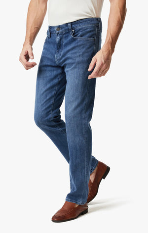A man wears 34 HERITAGE CHARISMA Relaxed Straight Leg Denim in Mid Urban Wash, a white shirt, and brown leather shoes, standing against a plain white background.