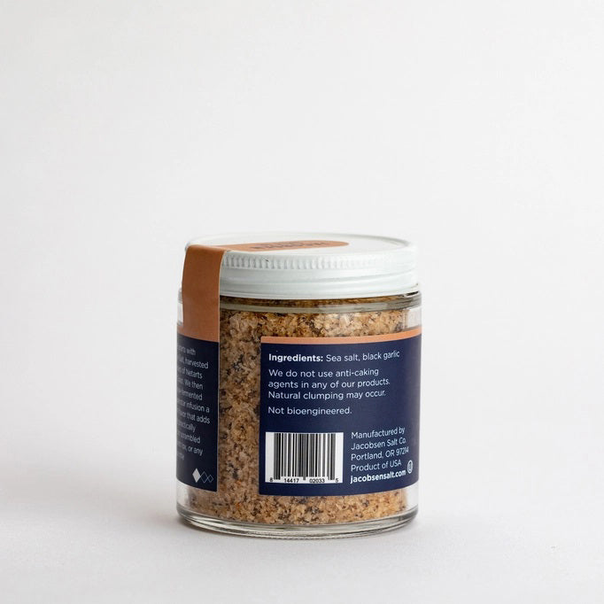 A jar of JACOBSEN SALT CO. INFUSED BLACK GARLIC SALT, featuring a black label and white lid, rests on a light beige surface against a plain light gray background.