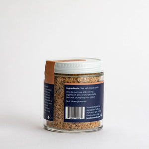 A jar of JACOBSEN SALT CO. INFUSED BLACK GARLIC SALT, featuring a black label and white lid, rests on a light beige surface against a plain light gray background.