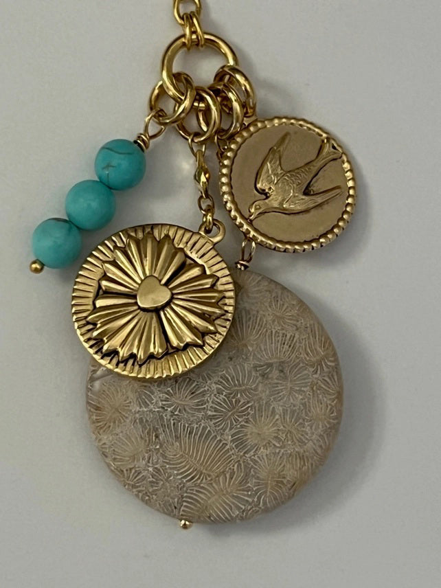 The SKYE NECKLACE by SANDRINE FRANCE STUDIO features a gold chain with a blue fish charm, turquoise beads, a bird-engraved gold coin, round gold charm, and a floral Fossil Coral pendant—a striking handmade jewelry piece.