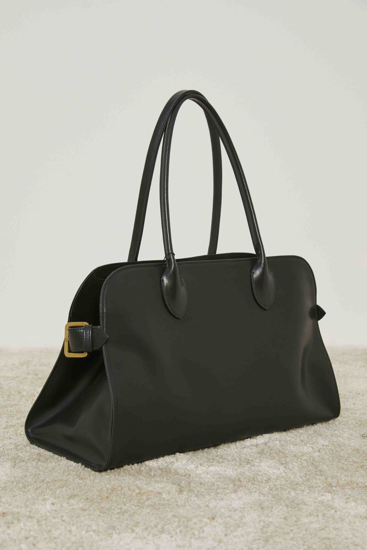 A person in loose cream pants and a matching sweater stands on carpet, holding the MEIMEIJ - BLACK LEATHER HANDBAG by its handles, adding a structured silhouette to the relaxed MEIMEIJ ensemble.