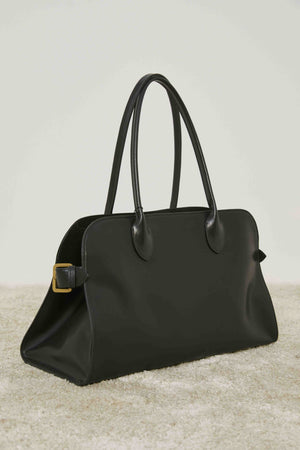 A person in loose cream pants and a matching sweater stands on carpet, holding the MEIMEIJ - BLACK LEATHER HANDBAG by its handles, adding a structured silhouette to the relaxed MEIMEIJ ensemble.