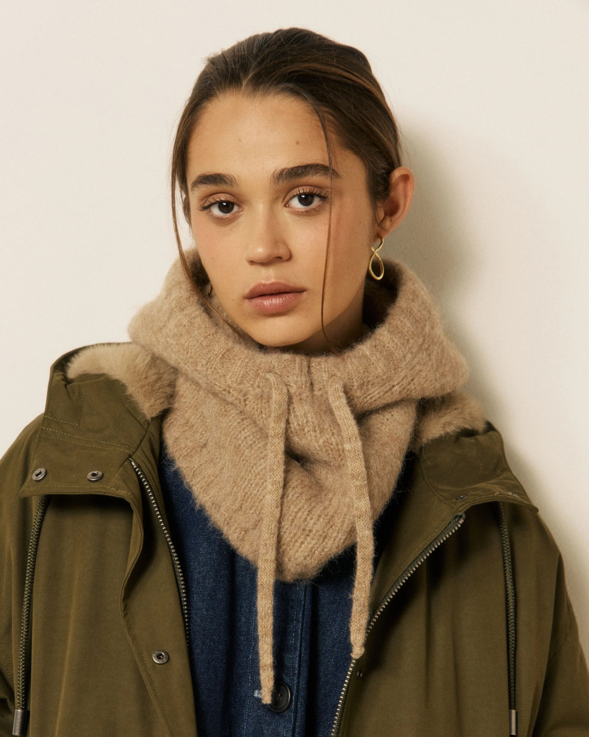 A woman with brown hair pulled back, wearing a green jacket, a navy top, and the HARTFORD - MEVA HOOD IN BEIGE by HARTFORD—a chunky tan knit merino wool scarf—along with a single gold hoop earring, stands against a plain off-white background.