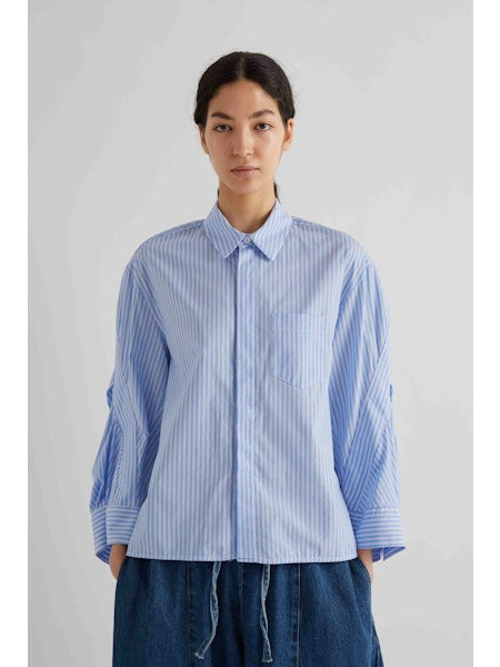 A woman with dark hair tied back models the MEIMEIJ STRIPE COTTON SHIRT—an oversized light blue and white striped cotton top with wide sleeves—paired with blue pants, standing against a plain light gray background.