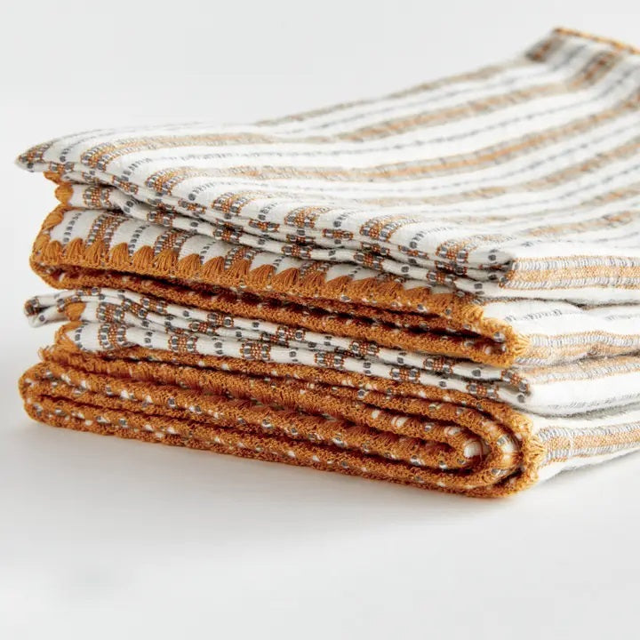 Elevate your table linens collection with the NAPA HOME AND GARDEN - SUTTON DINNER NAPKIN. This folded cotton napkin, featuring brown, white, and gray vertical stripes accented by decorative brown stitching along the edges, offers a stylish soft ochre touch—perfect for bringing both elegance and versatility to any dining setting. Displayed against a crisp white background, it’s a standout piece from NAPA HOME AND GARDEN.