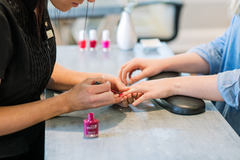A nail technician applying pink nail polish to a client's fingernails at a manicure station, with several bottles of nail polish and a small vase visible in the background.