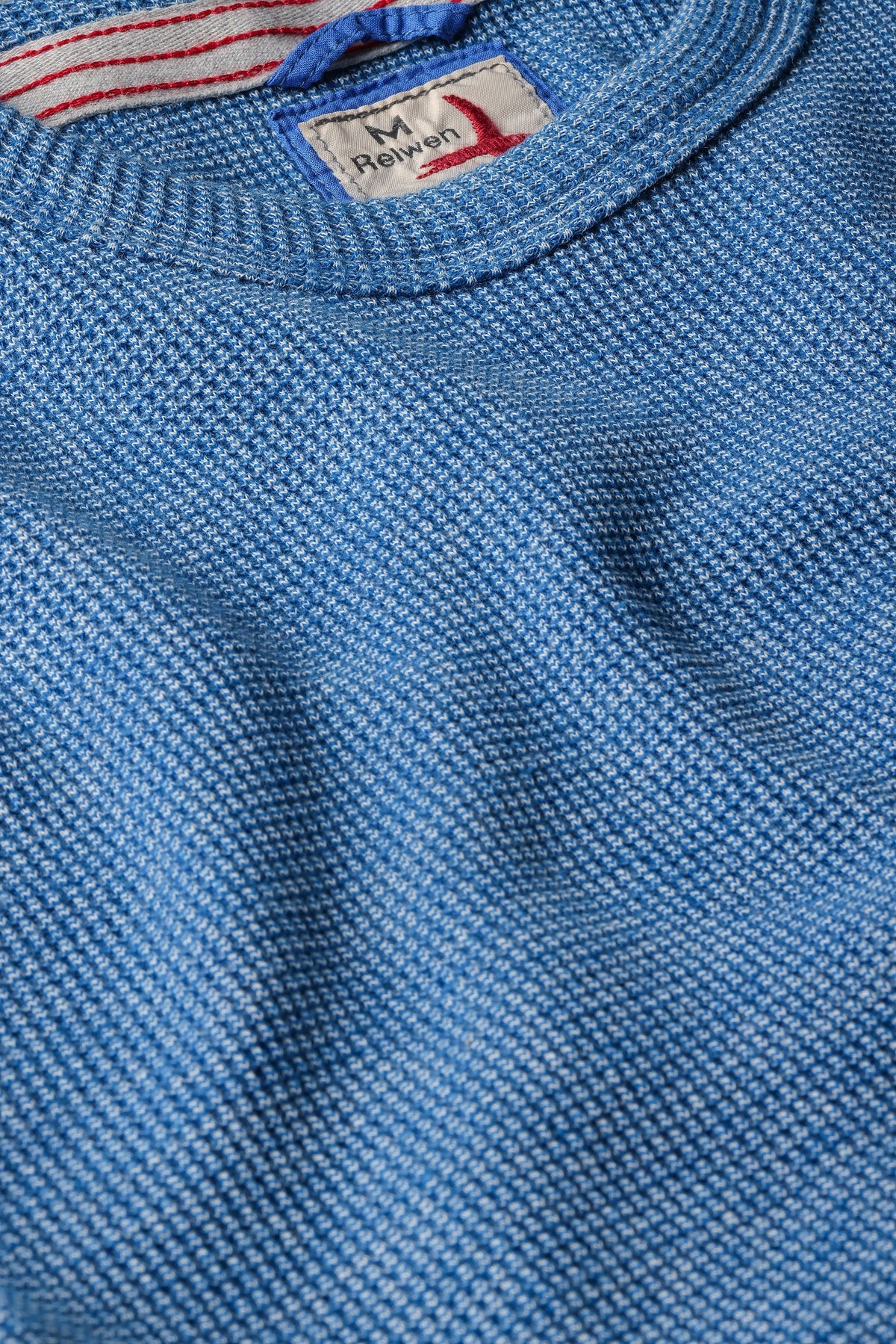 Close-up of the RELWEN - BI-WAFFLE CREW IN FRENCH BLUE, a textured light blue sweater with a vintage outdoor shirt vibe, featuring a visible collar and an inner neckline label showcasing text alongside a red and white stripe design.