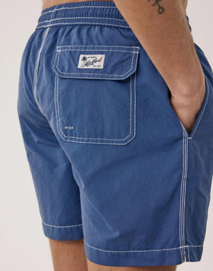 A shirtless person models the HARTFORD - NYLON SWIM TRUNKS WITH TRAVEL POUCH IN DENIM by HARTFORD against a plain light background, with tattoos visible on their arms and legs and one hand casually in a pocket.