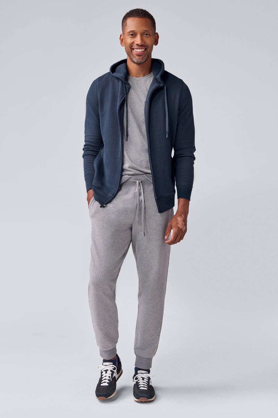 A man wears the MACK WELDON Ace Full-Zip Sweatshirt in True Navy (Bird's Eye) with light gray pants, standing against a plain light gray background and looking slightly to his left with a neutral expression.