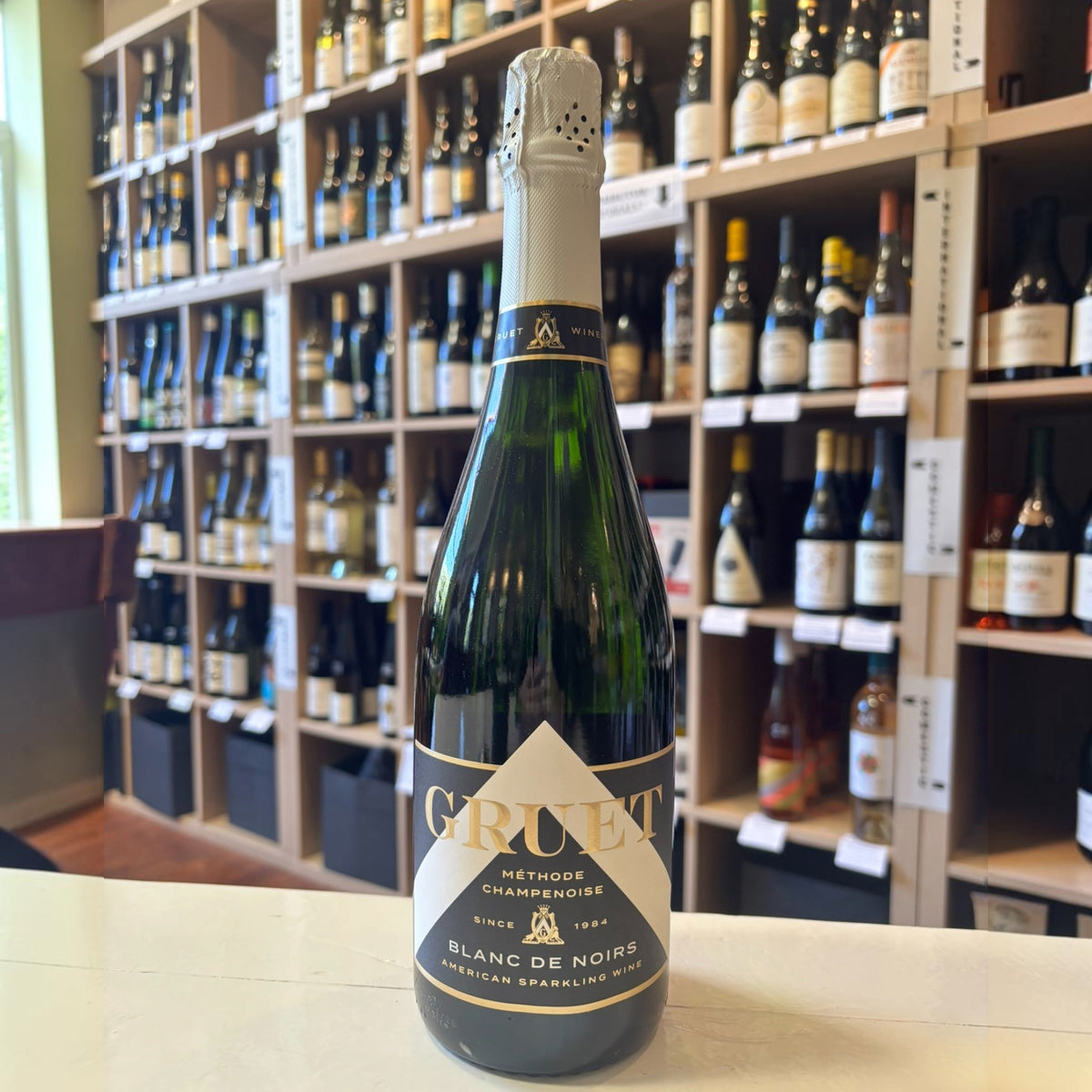 A bottle of Gruet Blanc de Noirs sparkling wine from New Mexico, crafted using the Champagne method, has a green glass body, beige foil on the neck, and a white label with black text for Wine: Empire Distributing.