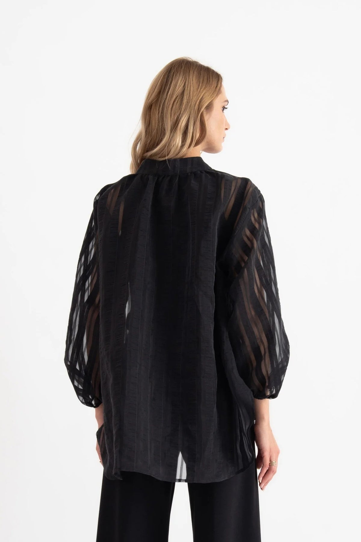 A woman stands in a bright room, looking at the camera with a neutral expression. She is wearing the VICARIO CINQUE SEAN SHEER STRIPE SHIRT, a black sheer long-sleeve blouse crafted from lightweight fabric with a band collar by VICARIO CINQUE, paired with black wide-leg pants and black sandals.