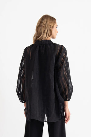 A woman stands in a bright room, looking at the camera with a neutral expression. She is wearing the VICARIO CINQUE SEAN SHEER STRIPE SHIRT, a black sheer long-sleeve blouse crafted from lightweight fabric with a band collar by VICARIO CINQUE, paired with black wide-leg pants and black sandals.