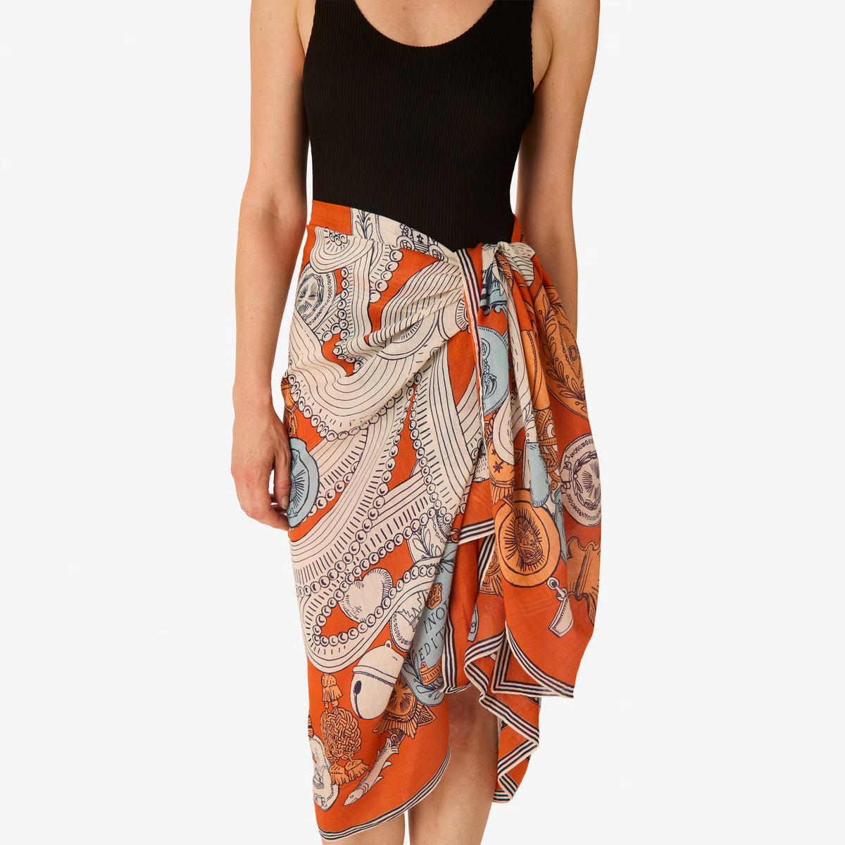 A woman with light skin and brown hair tied back wears a black outfit and the INOUI Editons Octopus Scarf in orange by INOUI, featuring intricate hand-printed patterns in orange, blue, and white draped over her shoulders.