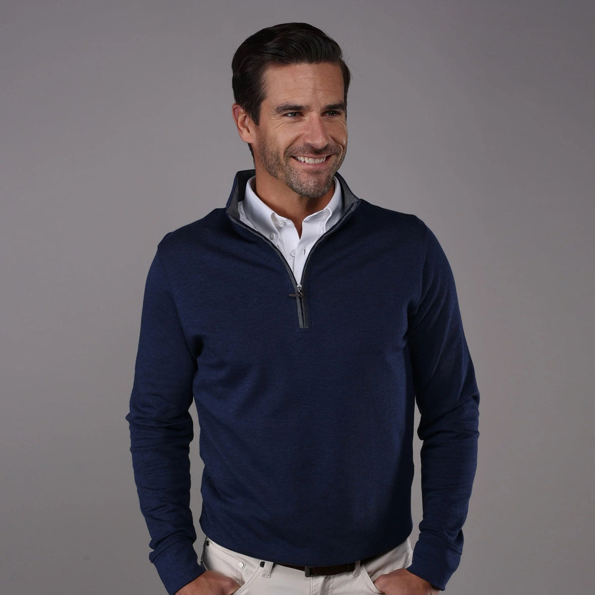 A man with short brown hair and a beard smiles while wearing the COLLARS AND CO - Heather Cotton Cloud 1/4 Zip Sweater in Navy with Grey Trim over a white collared shirt and cream pants, standing against a plain gray background.